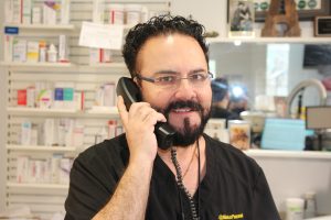A man with dark hair, beard, and glasses is smiling while talking on a corded phone in a pharmacy setting, surrounded by shelves with medication boxes.