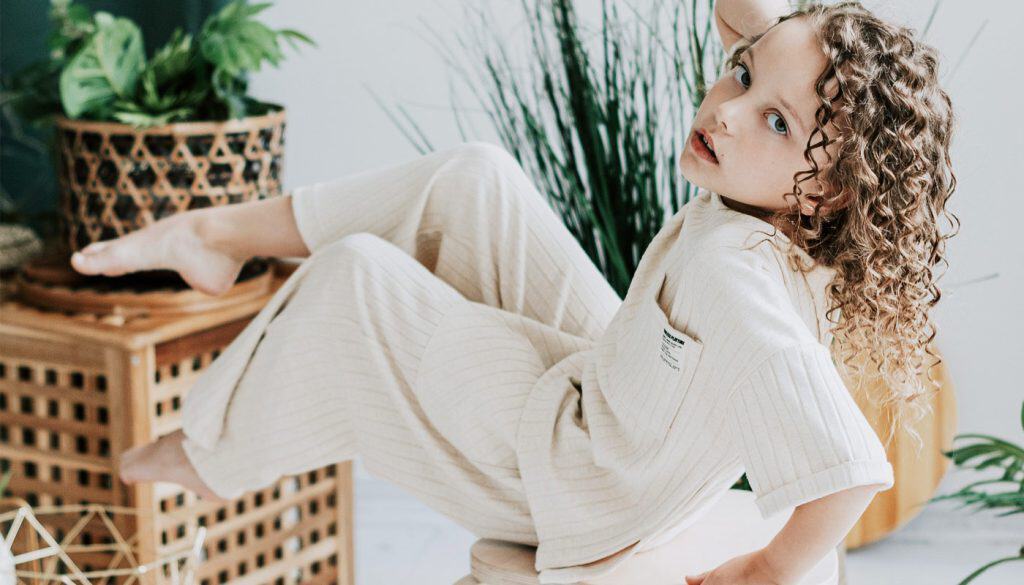 A child with curly hair poses playfully reclining on a stool in a light-colored outfit. Nearby, lush green plants and a wooden basket with geometric patterns create a cozy atmosphere akin to the welcoming vibe of the best pharmacy Toronto delights in offering.