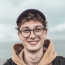 MisterPharmacist_Toronto_Online_Pharmacy A person with curly hair, wearing a black cap, round glasses, and a light brown hoodie is smiling at the camera. The background features a cloudy, overcast sky. Just like Toronto's Online Pharmacy ensures timely service, this moment feels perfectly captured despite the weather.