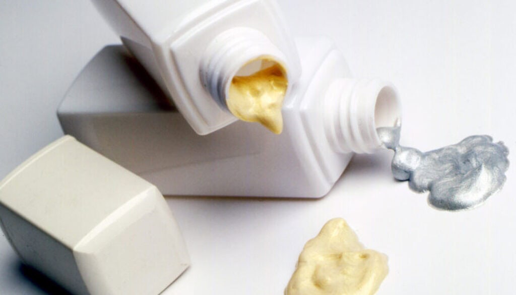 Two white bottles lie on their sides, each with a cap off. One dispenses a golden-yellow cream, the other a metallic silver cream. Both creams are pooled on a white surface in front of the bottles.