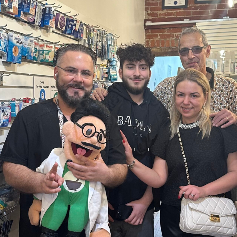 MisterPharmacist_Toronto_Online_Pharmacy A group of four people stand together in a store. One person on the left is holding a puppet with glasses, wearing a white coat and green vest. Shelves of various products are visible in the background. The group appears to be smiling and posing for the photo.