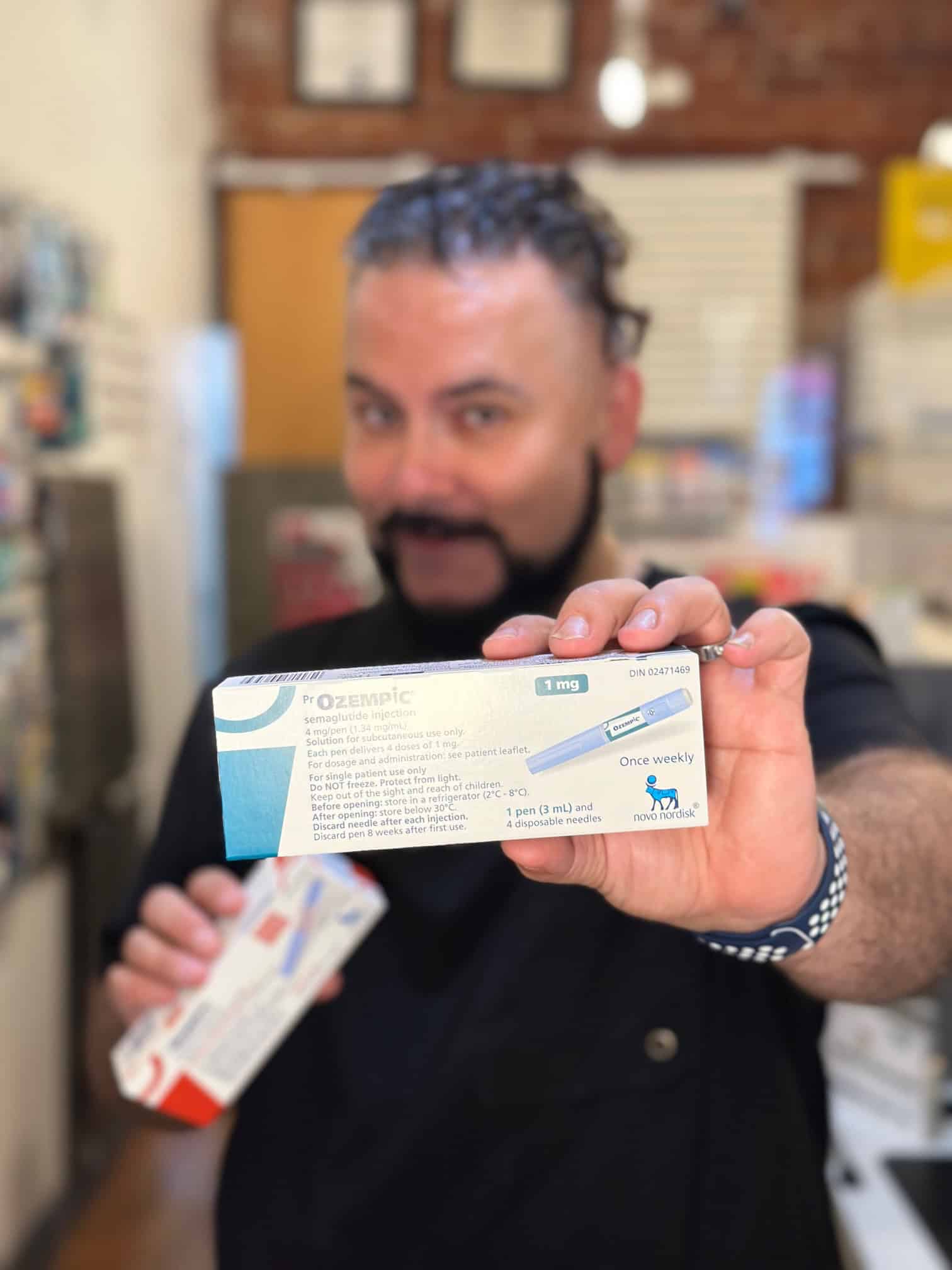 MisterPharmacist_Toronto_Online_Pharmacy A person with a beard and dark, curly hair, wearing a black shirt, holds a box of Ozempic medication in a blurred background of a pharmacy or medical store. The box is focused and clearly visible, showing dosage information and branding.