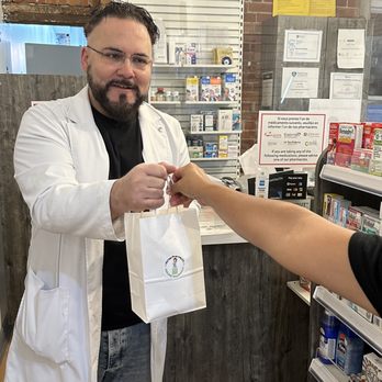 MisterPharmacist_Toronto_Online_Pharmacy A pharmacist in a white coat hands over a small paper bag to a customer across the counter in TorontoOnlinePharmacy. Shelves with various products are visible in the background, highlighting the fast prescription delivery service.