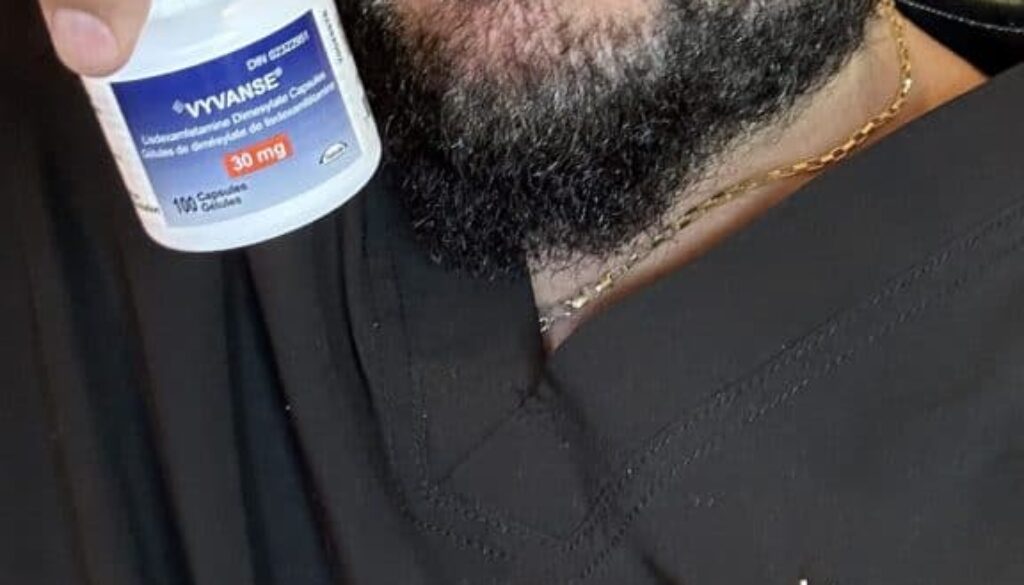 A man wearing dark scrubs labeled "Mister Ph" holds up a bottle of Vyvanse 30 mg capsules close to his face, looking directly at the camera.