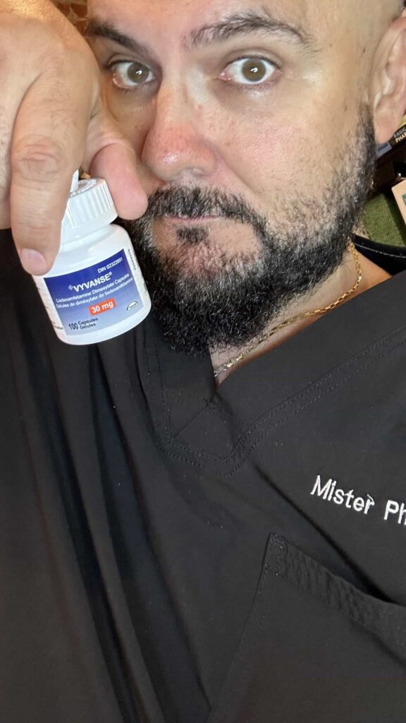 A man wearing dark scrubs labeled "Mister Ph" holds up a bottle of Vyvanse 30 mg capsules close to his face, looking directly at the camera.