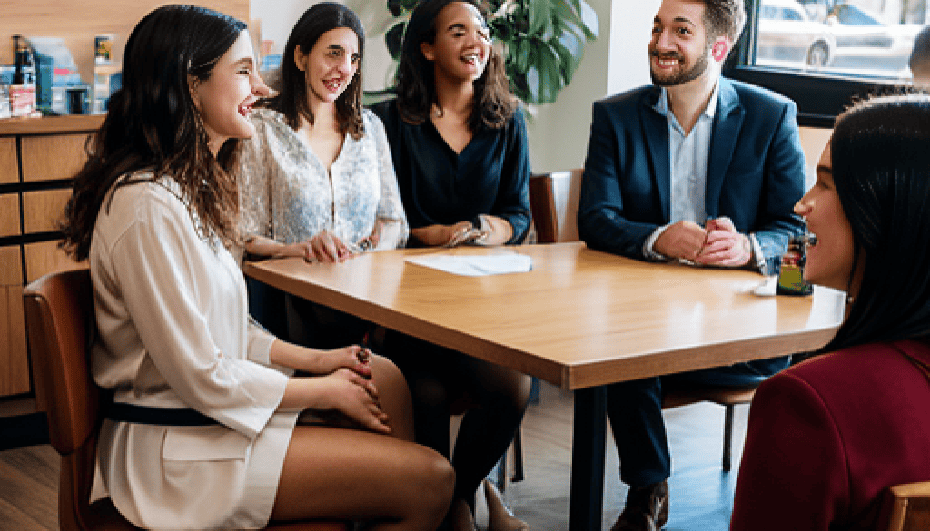 MisterPharmacist_Toronto_Online_Pharmacy A group of five people, three women and two men, sit around a wooden table in a bright office. They are smiling and engaged in conversation. Shelves with various items and a large window are in the background.