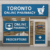 Storefront of a Toronto online pharmacy with blue signs displaying "Online Pharmacy" and "Prescriptions," a mortar and pestle icon, and an "Open" sign on the door. Shelves with products are visible inside.