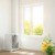 Bright indoor scene with air purifier by window closed against pollen; sunlight filters in, showing blooming flowers and clear sky outside.