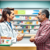 A pharmacist consulting with a middle-aged man at a pharmacy counter, surrounded by medication shelves and health brochures.