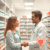 Bright pharmacy interior with shelves of medications, a pharmacist assisting a patient, and subtle urinary tract health icons visible.