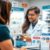 A pharmacist in a white coat smiles while assisting a customer at the pharmacy counter, with ear care products and informational displays about swimmer's ear arranged in front of them. Pharmacist consulting patient about ear care with ear drop products and swimmer's ear brochures on the pharmacy counter.