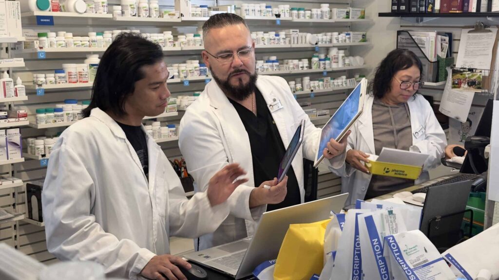 Three pharmacists in white lab coats collaborate in a Toronto online pharmacy. They are reviewing documents and using a laptop. Shelves filled with medication bottles are visible in the background, embodying the essence of the best pharmacy Toronto has to offer.