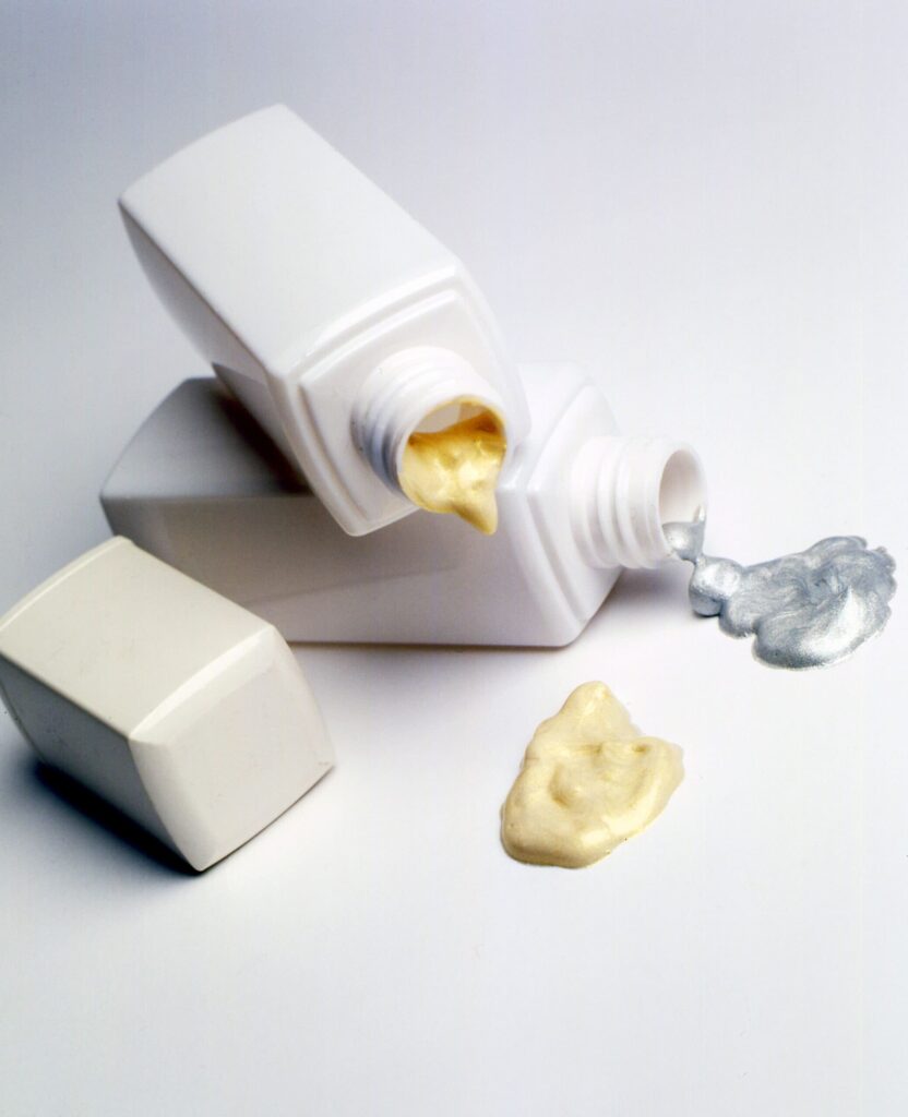 Two white bottles lie on their sides, each with a cap off. One dispenses a golden-yellow cream, the other a metallic silver cream. Both creams are pooled on a white surface in front of the bottles.